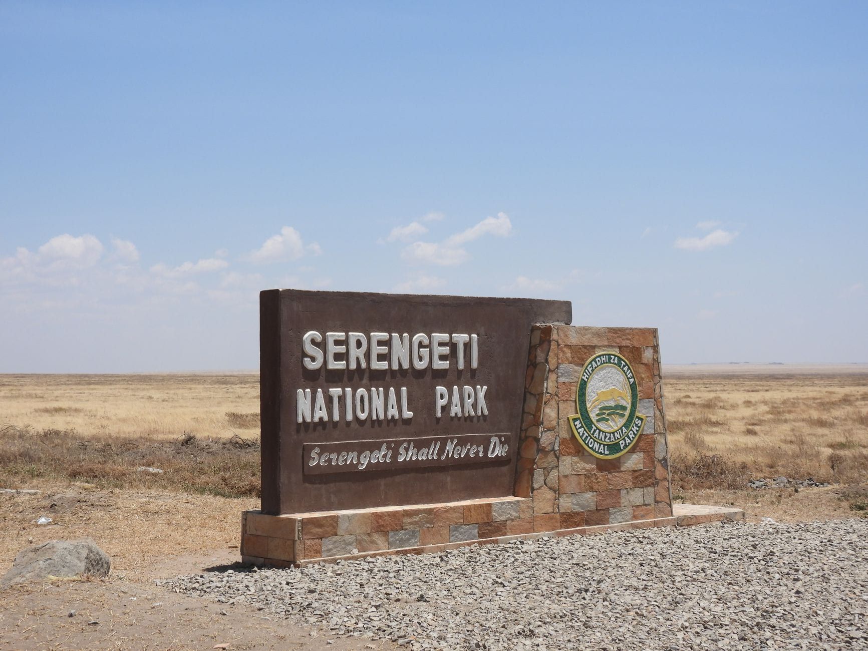 Ein Eingangsschild für den Serengeti-Nationalpark steht vor weitläufigem, trockenem Grasland unter einem teils bewölkten Himmel.